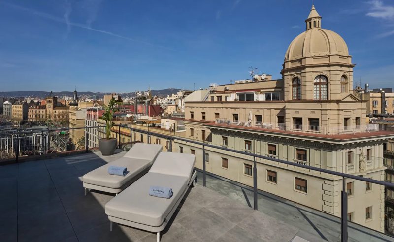 Terraço do hotel com espreguiçadeiras e vista dos telhados de Barcelona, com uma cúpula ao lado