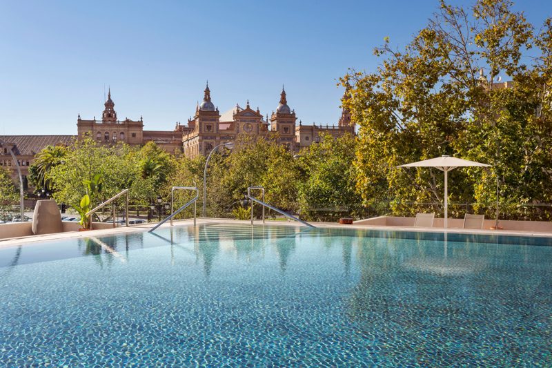 Piscina do hotel entre árvores, com as torres da Plaza de España ao fundo